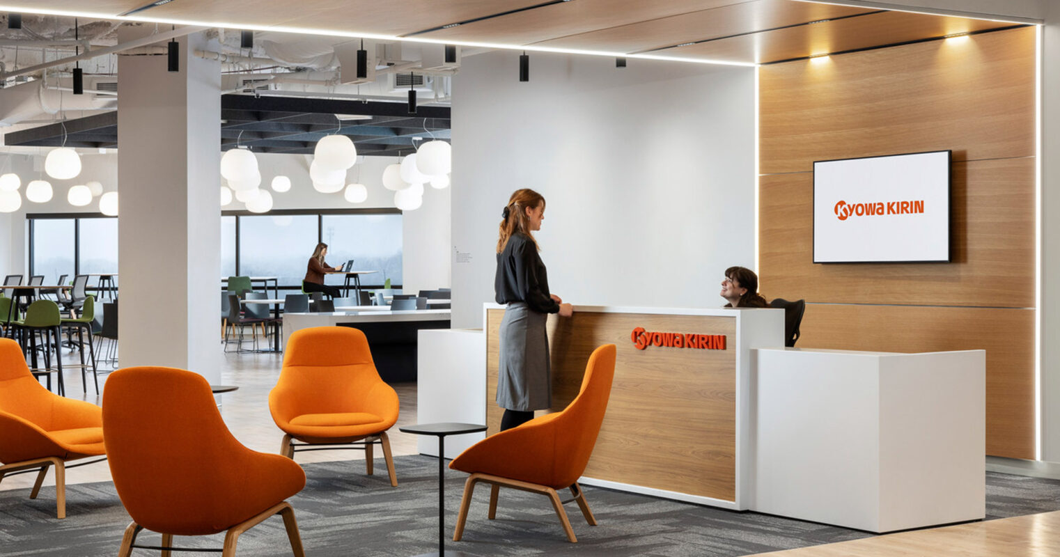 Modern office lobby featuring a sleek reception desk with a lit company sign, vibrant orange chairs adding a pop of color, and pendant lighting illuminating the open, airy space.