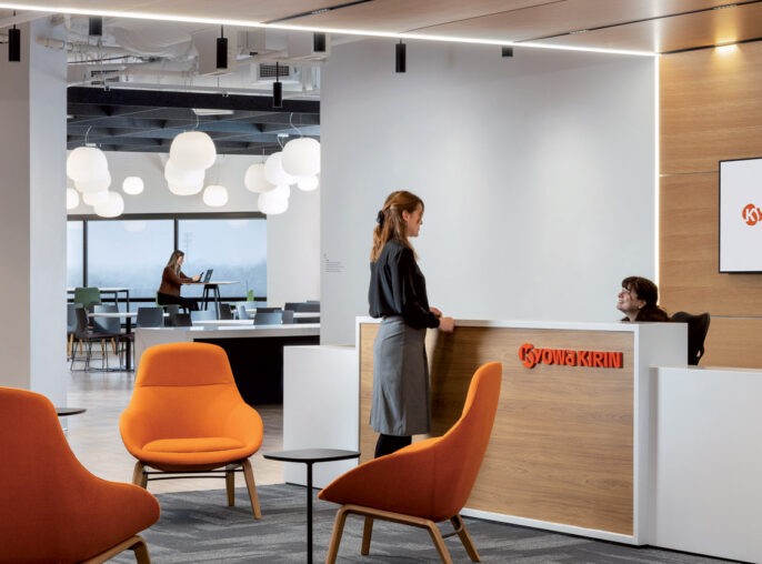 Modern office lobby featuring a sleek reception desk with a lit company sign, vibrant orange chairs adding a pop of color, and pendant lighting illuminating the open, airy space.