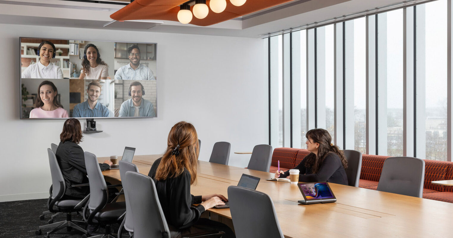Modern conference room featuring a large wooden table, surrounded by ergonomic chairs and paneled with floor-to-ceiling windows. Overhead, an array of circular pendant lights adds warmth. On the wall, a video conference call is in progress, blending in-person and virtual collaboration.