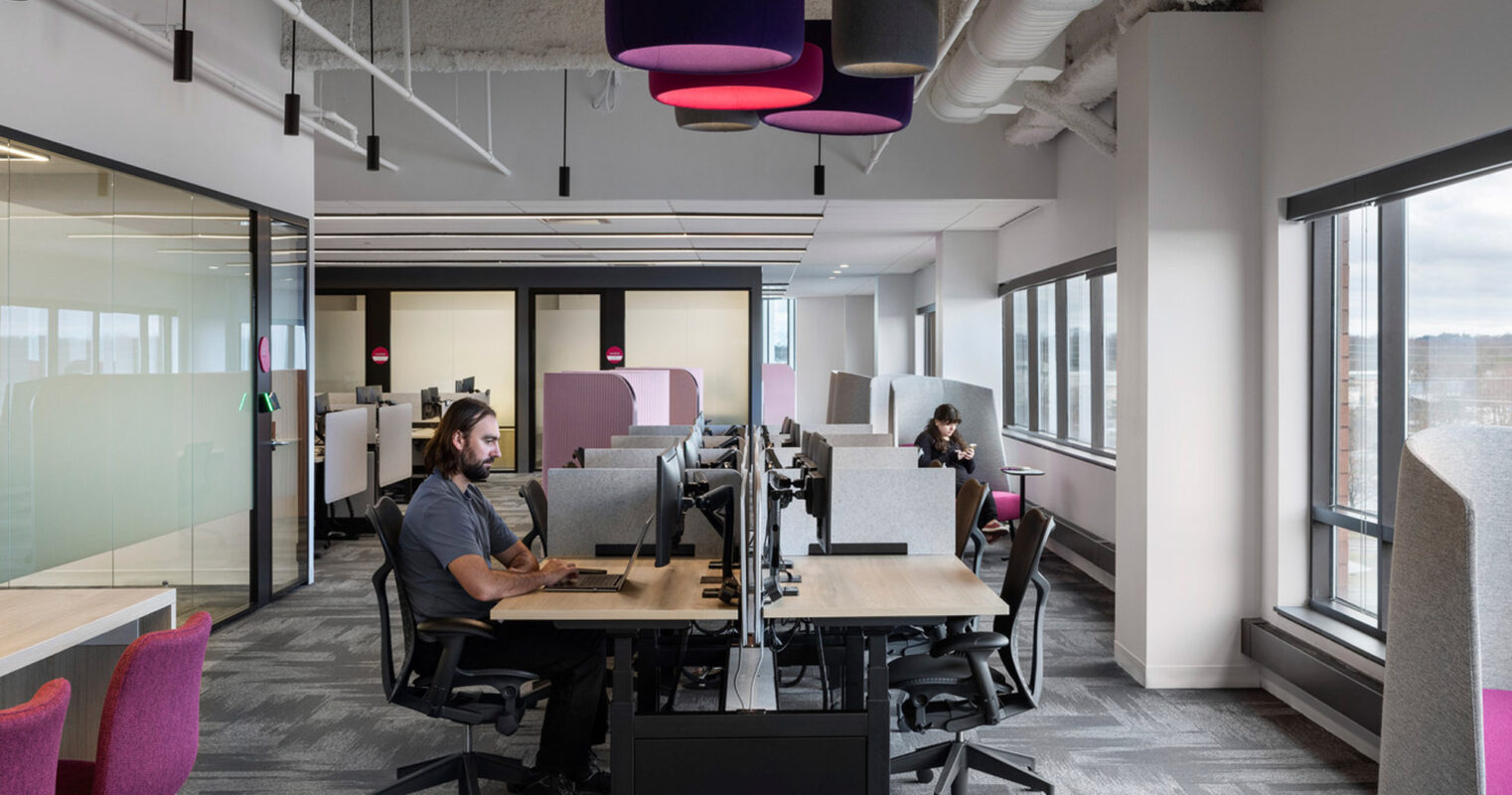 Modern office interior showcasing an open-plan layout with individual workstations, ergonomic chairs, and vibrant fuchsia accents from overhead lighting fixtures and seating. Clear glass partitions maintain openness, while exposed ceiling ducts add an industrial touch juxtaposed with the carpeted floors for a contemporary workspace design.