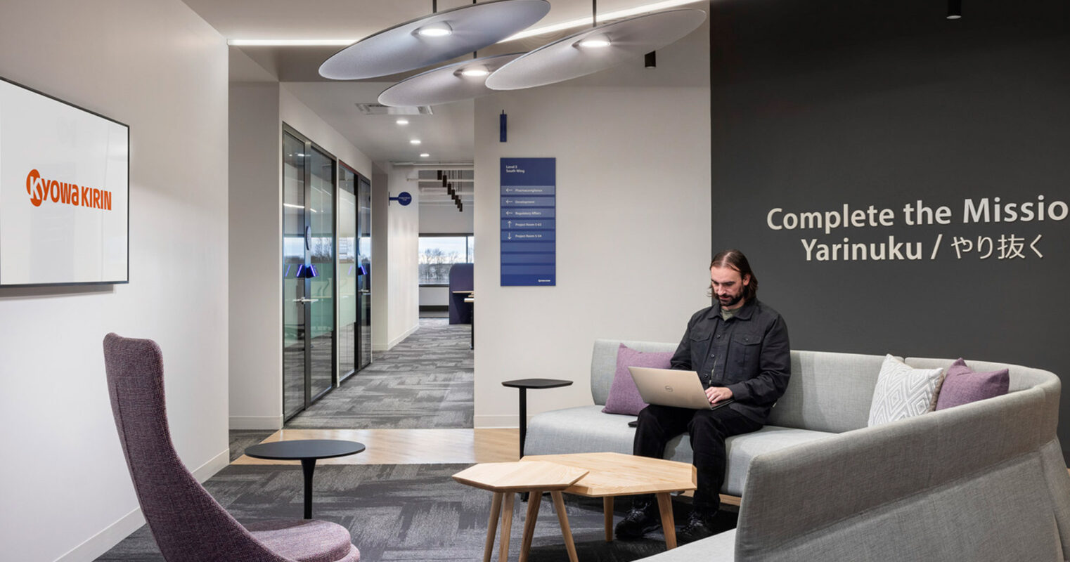 Modern office lounge with minimalist furnishings including a gray sofa, complemented by purple armchairs, and round wooden tables. A man works on a laptop, under unique circular lighting fixtures, near a wall with bold lettering, creating a functional yet stylish workspace.