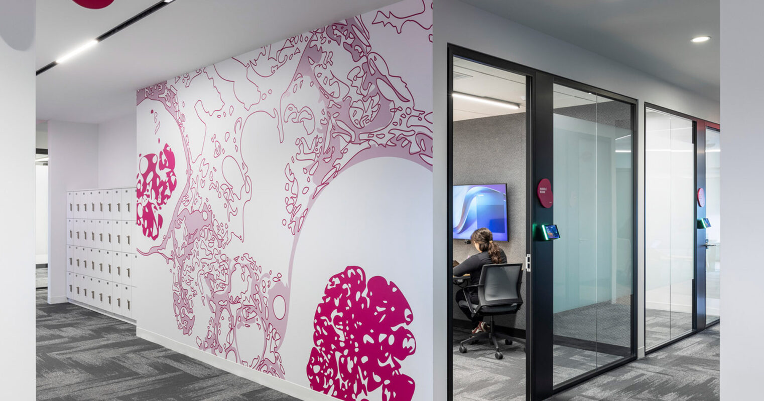 An office foyer featuring a striking magenta and white abstract wall graphic that contrasts with the grayscale carpeted floor. Glass doors with magenta decals lead to a workspace where an individual is seated, enhancing the space's contemporary and creative appeal.