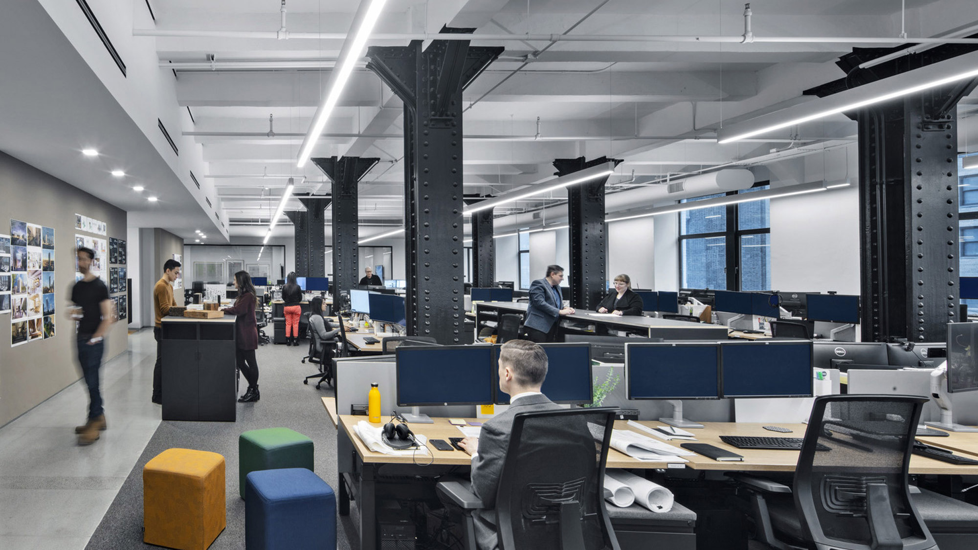Modern open-concept office space with exposed black steel columns, ergonomically designed workstations, and pendant lighting. Neutral color palette highlighted by natural light and accentuated by pops of yellow and blue in the seating areas. A collaborative environment is suggested by communal workspaces and a pin-up board-clad corridor.