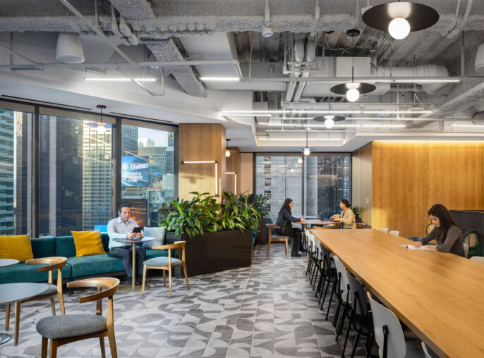 A modern office break room featuring a geometric-patterned floor, various seating options including bar-height tables and plush couches, accented by vibrant furnishings and ample greenery, bordered by floor-to-ceiling windows overlooking an urban landscape.