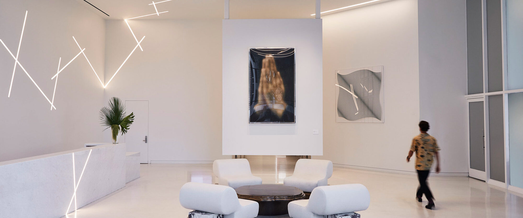 Modern interior design showcasing a minimalist aesthetic with stark white walls, polished concrete floors, and abstract ceiling lighting. Central seating area features sleek armchairs around a dark wooden table, complemented by large-scale contemporary artwork on the walls.