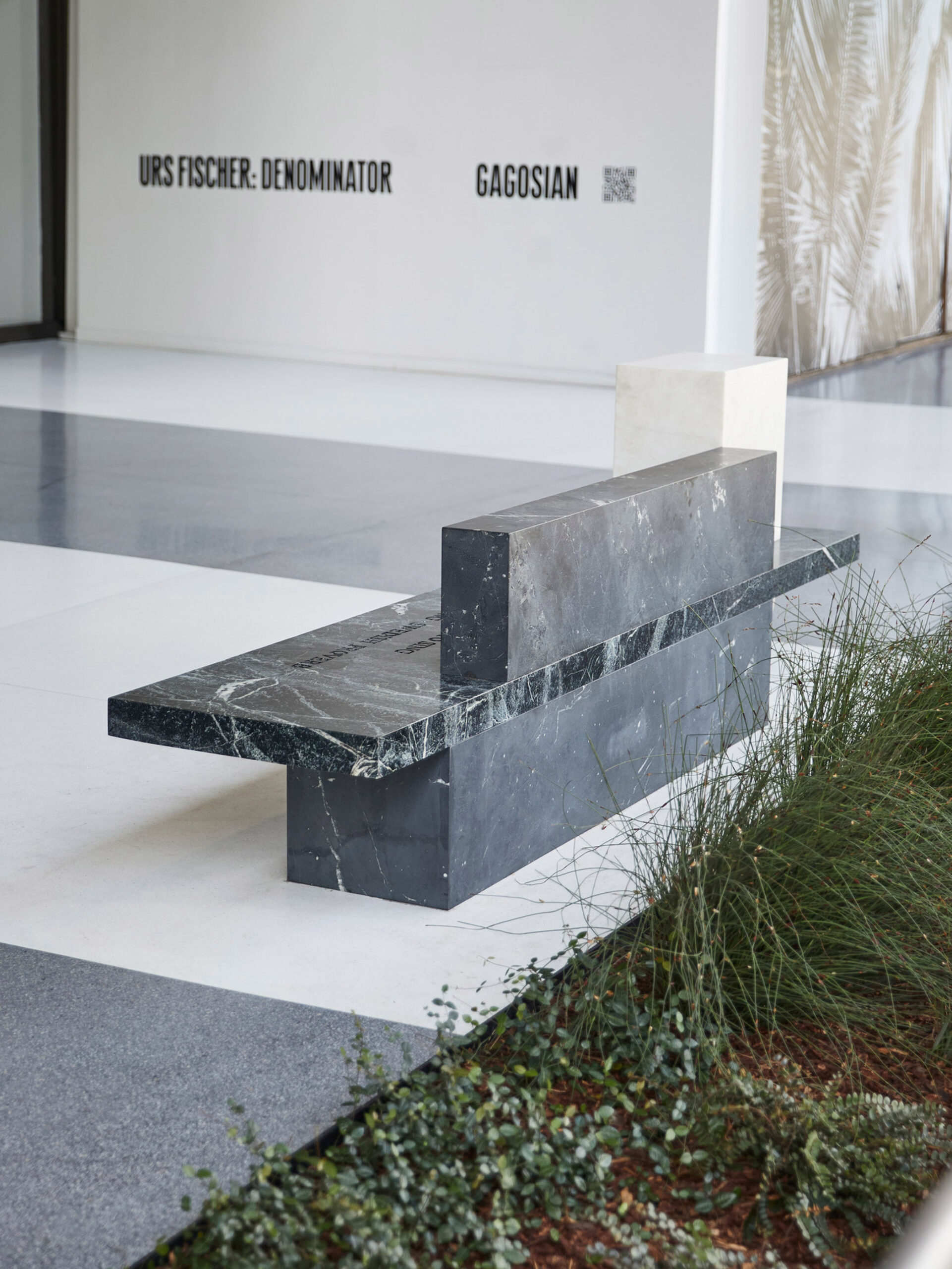 Minimalist marble bench with a contrasting geometric design, positioned in a spacious gallery with polished concrete floors. The simple, sleek lines of the bench provide a modern contrast against the organic textures of the adjacent greenery.