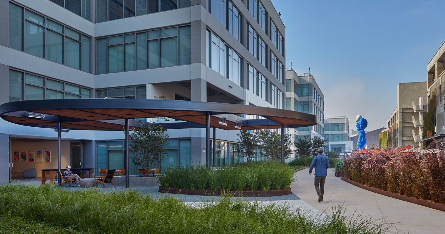 Modern urban landscape featuring an outdoor communal area with sleek, overhanging canopies, wooden deck chairs, and lush greenery, nestled between contemporary multi-story buildings.