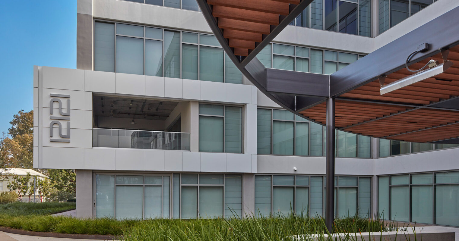 Modern architectural facade featuring a juxtaposition of materials with reflective glass windows, sleek metal paneling, and warm-toned wooden accents under a clear blue sky. Landscaping includes lush, green ornamental grasses complementing the building's linear design elements.