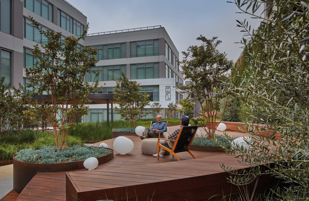 Two people sitting on a wooden deck in a urban, landscaped garden surrounded by buildings.