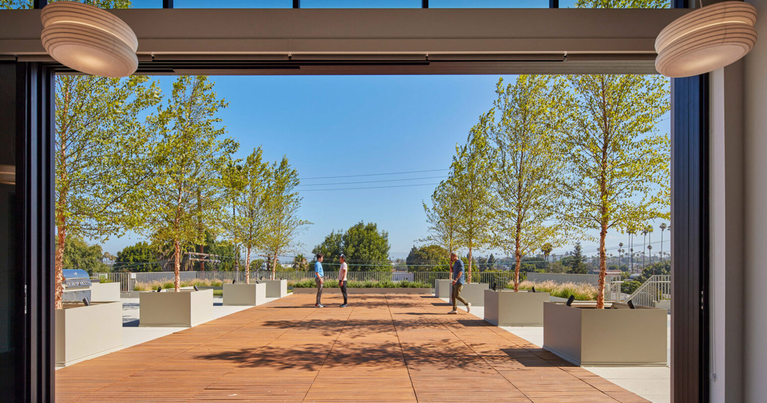 Spacious room with floor-to-ceiling retractable glass wall, seamlessly connecting the indoor polished concrete flooring to an outdoor wooden deck. Overhead cylindrical lighting fixtures complement the modern aesthetic, framing the view of the tranquil exterior landscape and pedestrians.