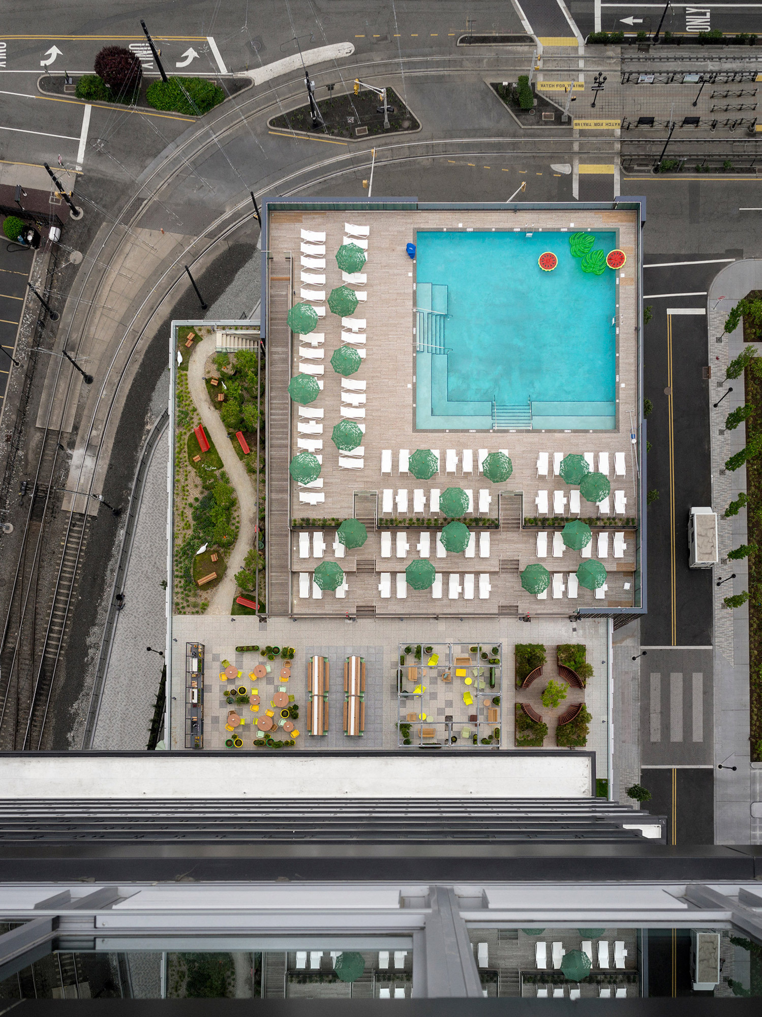 Bird's-eye view captures a rooftop pool with geometric lounging areas, surrounded by a perimeter garden. The juxtaposition of the aquatic blue against the urban grays highlights a well-planned oasis amidst the city's structured lines.