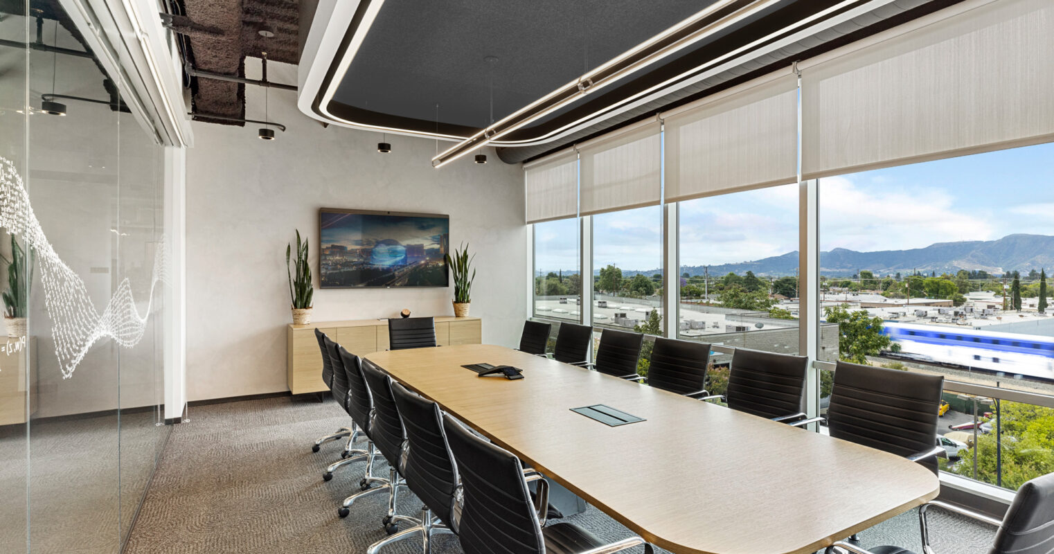 Modern conference room featuring a long rectangular table with black swivel chairs, floor-to-ceiling windows offering mountain views, a textured ceiling with integrated lighting, and an abstract wave-like glass partition.