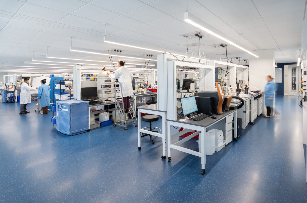 Modern laboratory space featuring streamlined workstations equipped with advanced technology. The open layout promotes collaboration, allowing researchers to interact freely. Bright overhead lighting and smooth, blue flooring enhance visibility and cleanliness, while laboratory equipment is neatly organized to create an efficient workflow. People in lab coats engage in various tasks.