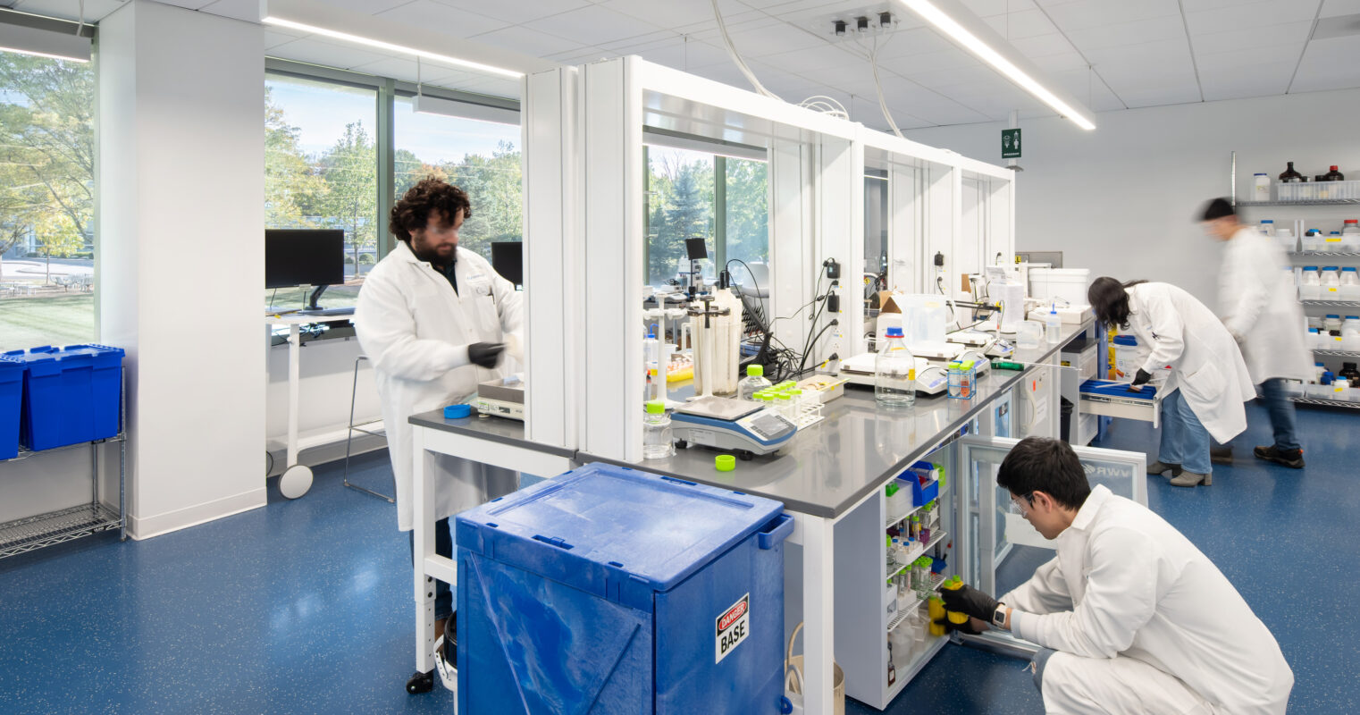 Bright, modern laboratory featuring spacious workstations equipped with laboratory equipment and supplies. Researchers in white lab coats actively engage in experiments, emphasizing collaboration. Large windows allow natural light, enhancing the workspace's functionality. Sleek designs and organized storage contribute to an efficient and professional research environment.