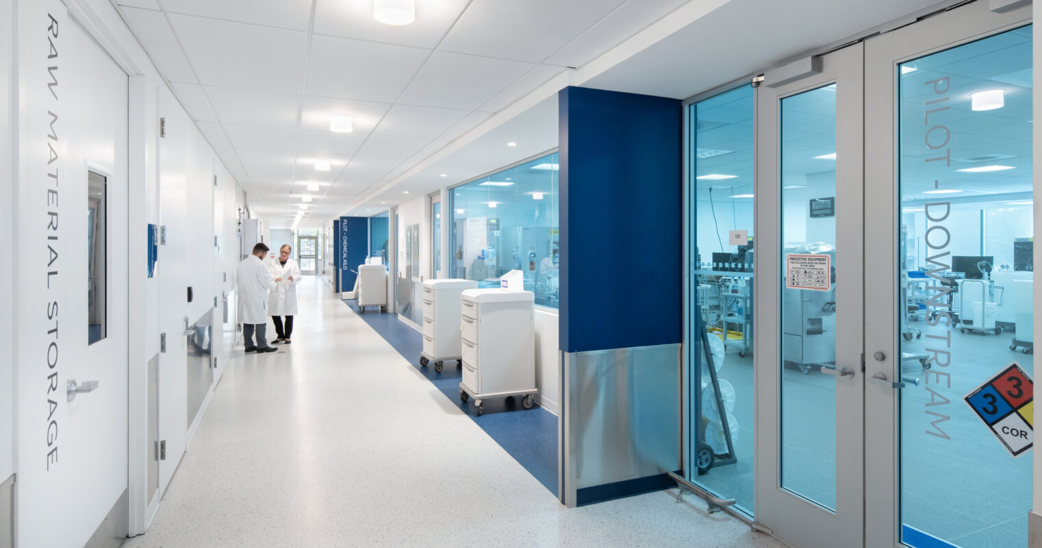 Bright, modern corridor in a healthcare facility featuring sleek white walls with blue accents. Large glass doors lead to sterile workspaces. Clean design elements include seamless flooring and overhead lighting, emphasizing functionality and space efficiency in an environment tailored for medical professionals and high standards of cleanliness.