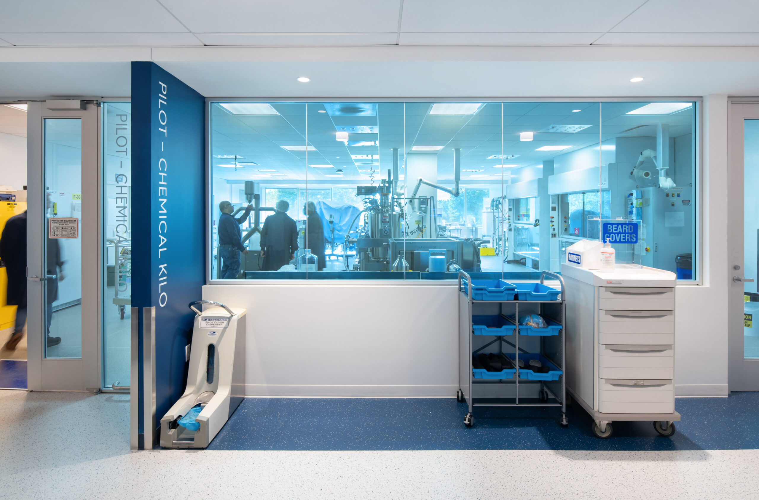 Modern laboratory interior featuring a bright, open design with large glass windows. The space showcases advanced equipment and a streamlined layout, emphasizing functionality and collaboration. A mobile cart with storage bins stands in the foreground, alongside modern lighting that enhances the sterile atmosphere.