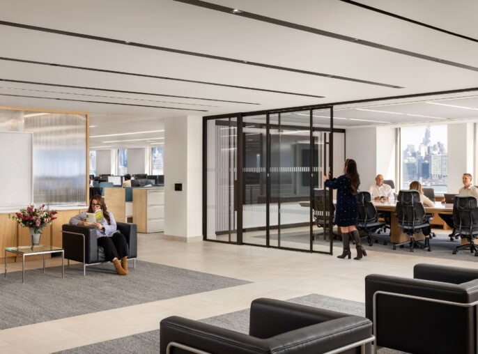 A modern open-plan office lobby features sleek black leather seating and a soft gray area rug, creating a welcoming atmosphere. Large glass partitions delineate a conference room, while natural light floods the space through expansive windows, highlighting the minimalist design and contemporary furnishings.
