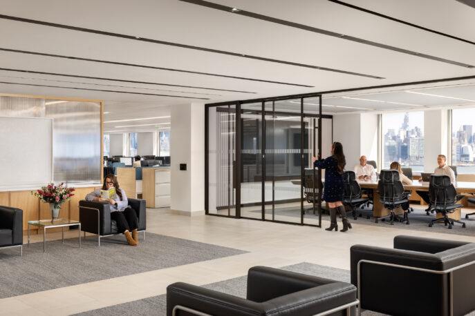 A modern open-plan office lobby features sleek black leather seating and a soft gray area rug, creating a welcoming atmosphere. Large glass partitions delineate a conference room, while natural light floods the space through expansive windows, highlighting the minimalist design and contemporary furnishings.