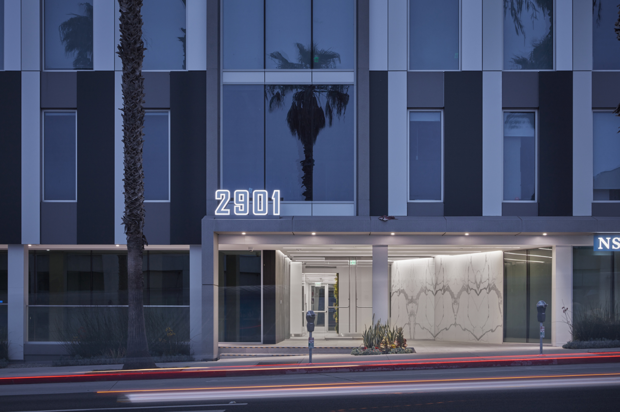 Modern architectural facade featuring vertical stripes in dark and light hues. The entrance is accentuated by illuminated numbers '2901' and sleek marble detailing, complementing the surrounding palm trees. A clean, minimalist aesthetic enhances the contemporary urban environment, inviting visitors with understated elegance.