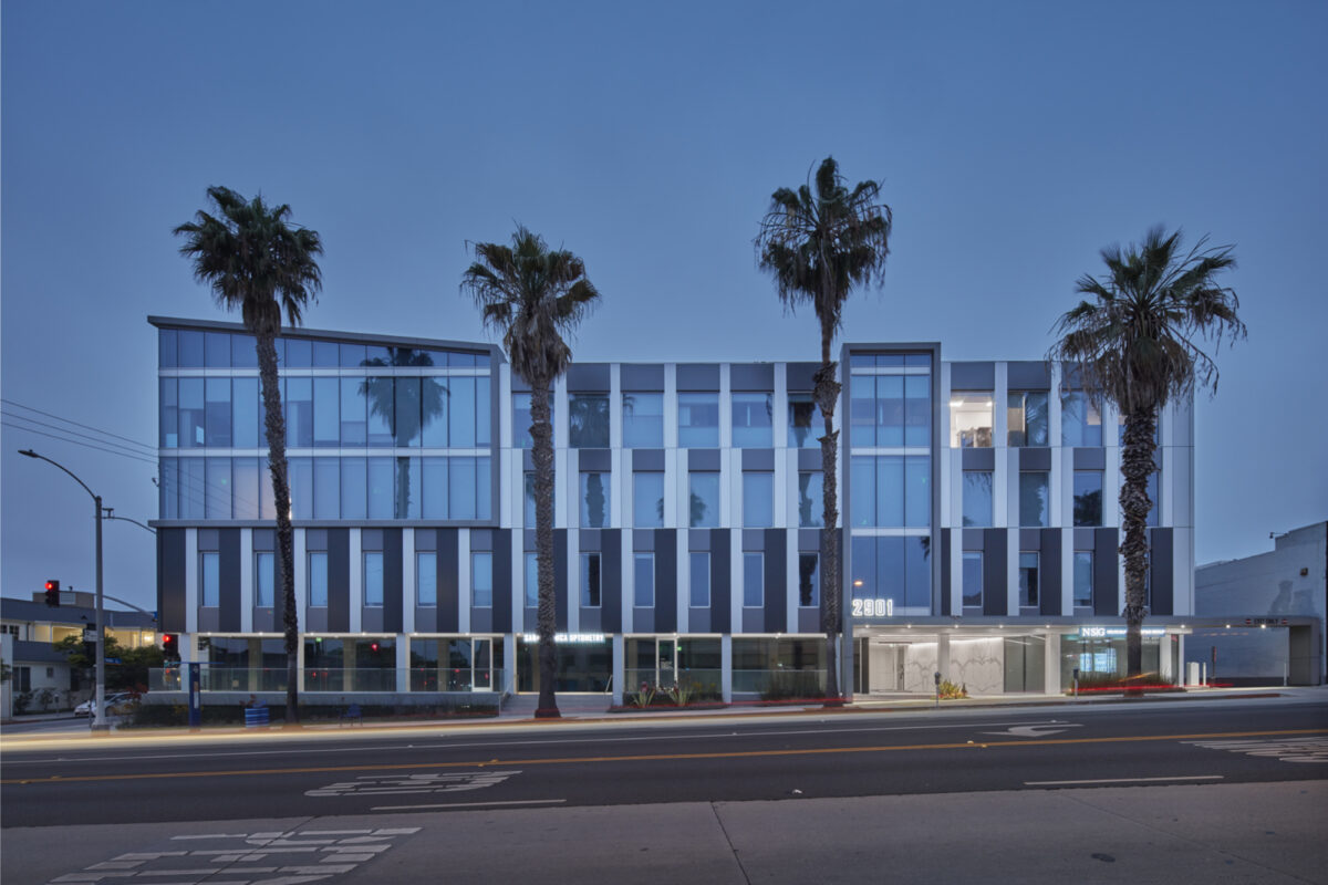Modern three-story commercial building featuring a sleek glass facade and vertical wooden slats. Tall palm trees flank the entrance, enhancing the tropical ambiance. The lighting at dusk illuminates the structure's contemporary lines and open spaces, creating a welcoming urban environment. Ideal for mixed-use purposes in a vibrant setting.