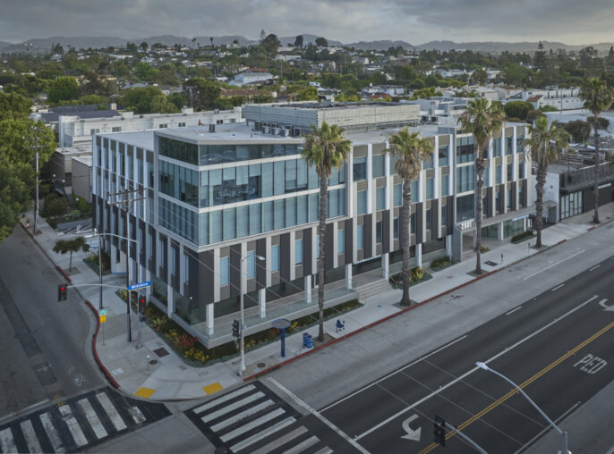 Modern commercial building featuring a sleek, geometric façade with alternating dark and light tones. Large glass windows reflect the surroundings, enhancing natural light. Palm trees line the street, contributing to a vibrant, urban atmosphere, while the structure’s design emphasizes functionality and contemporary architectural aesthetics.