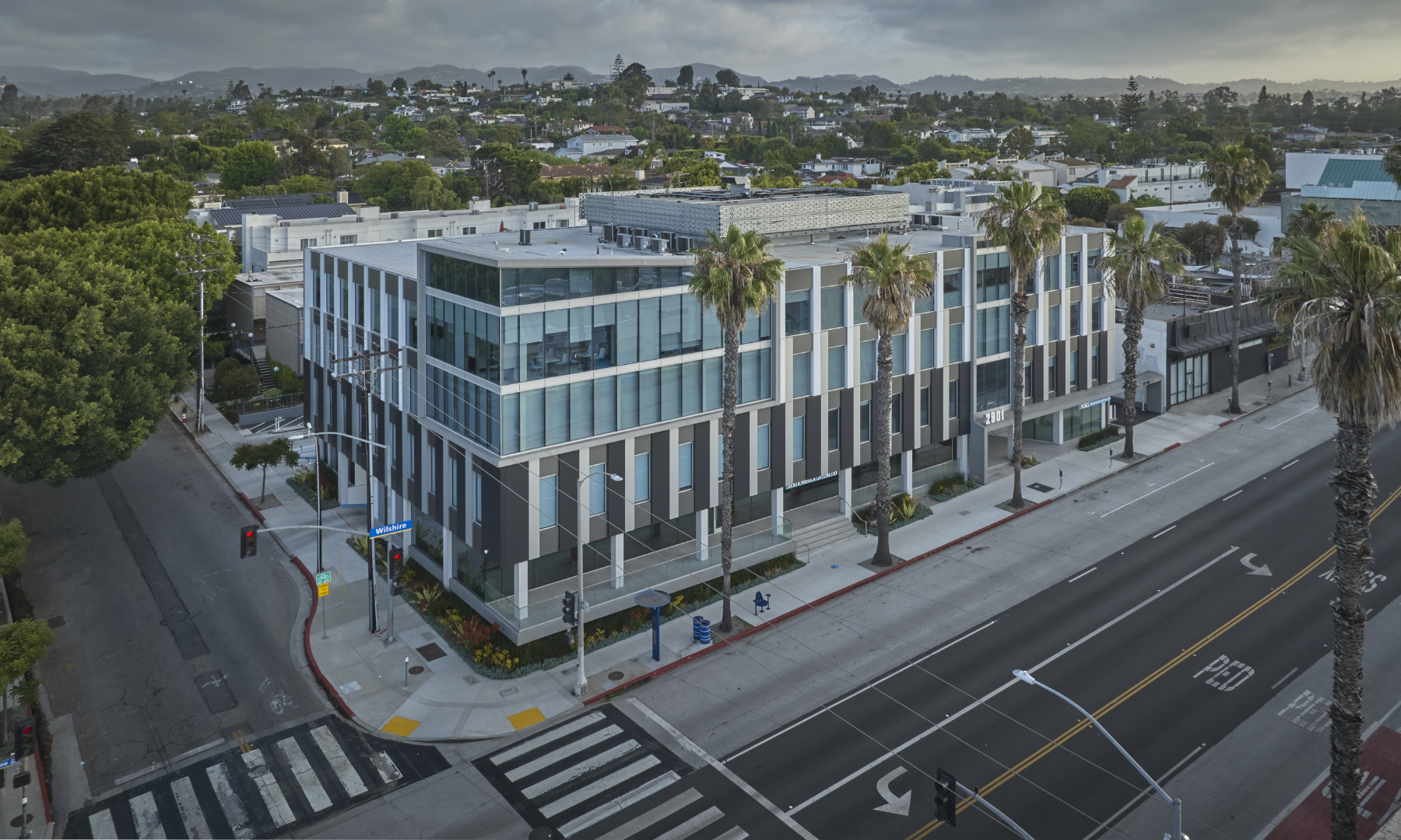 Modern commercial building featuring a sleek, geometric façade with alternating dark and light tones. Large glass windows reflect the surroundings, enhancing natural light. Palm trees line the street, contributing to a vibrant, urban atmosphere, while the structure’s design emphasizes functionality and contemporary architectural aesthetics.