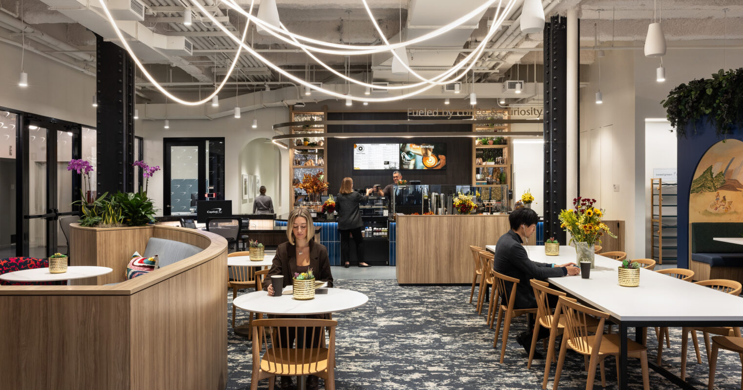 A modern, open-concept café features natural wood furnishings and a lively atmosphere. Dramatic, sculptural lighting casts soft shadows across the space. Guests enjoy the warm environment, surrounded by vibrant floral arrangements and a stylish beverage counter, while decorative elements add a unique touch to the overall design.
