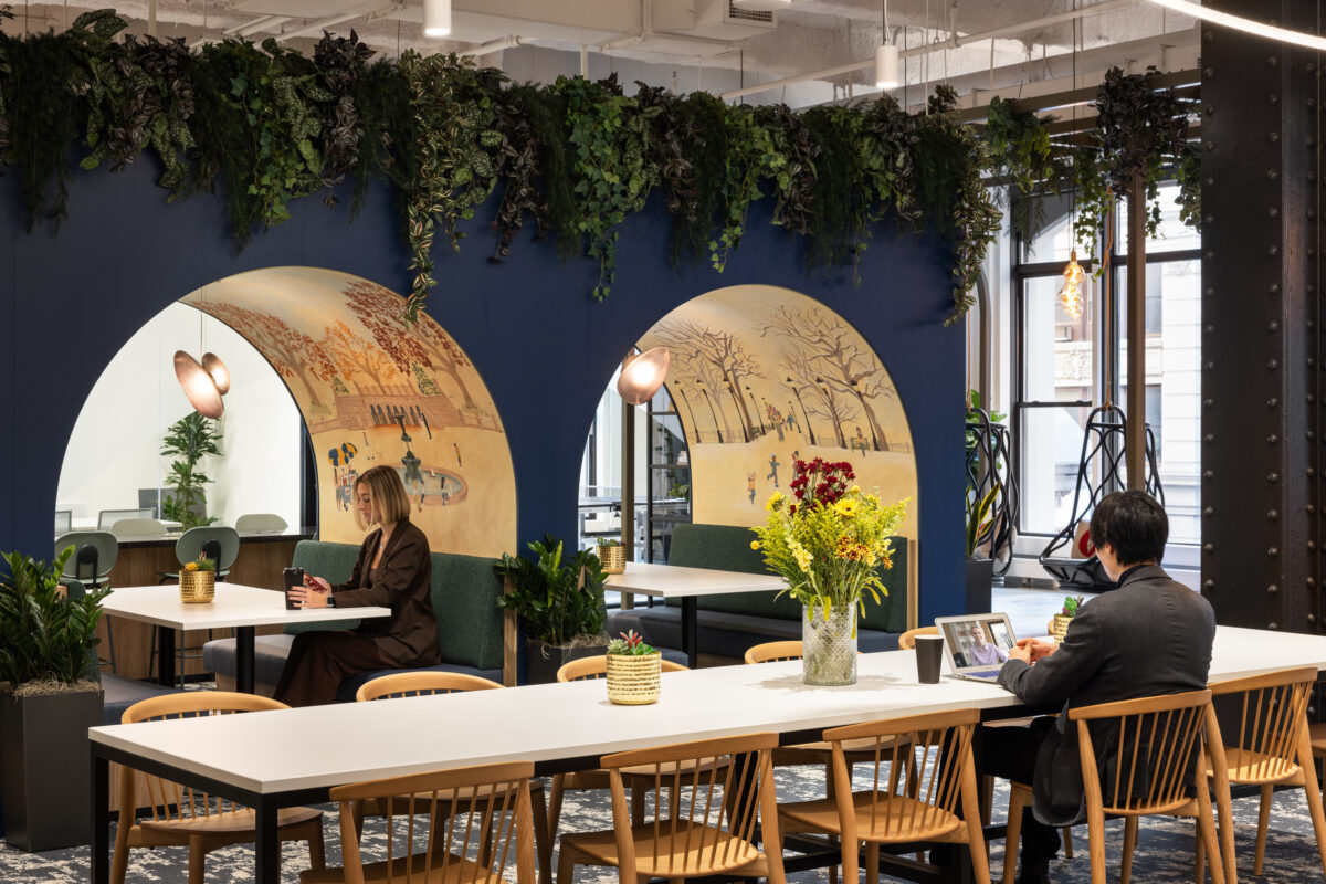 A modern office space features an inviting communal area with two large, rounded arches. Each arch showcases intricate mural art depicting serene landscapes, complemented by lush greenery overhead. Clean white tables contrast with natural wood chairs, while potted plants add a vibrant touch, creating a harmonious blend of work and relaxation.
