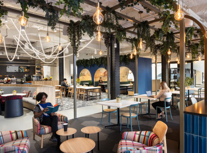 A modern co-working space featuring vibrant striped armchairs, natural greenery overhead, and artistic murals. The layout promotes collaboration, with open seating arrangements, communal tables, and individual workstations. Warm pendant lighting enhances the inviting atmosphere, making it a conducive environment for productivity and creativity.
