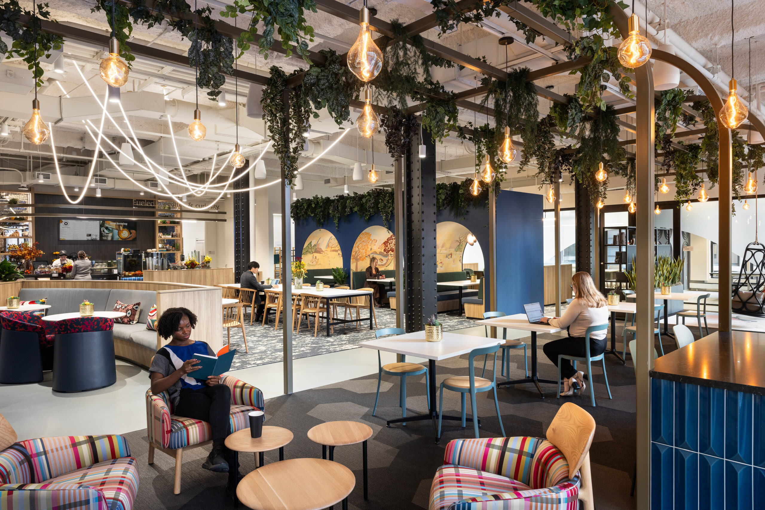 A modern co-working space featuring vibrant striped armchairs, natural greenery overhead, and artistic murals. The layout promotes collaboration, with open seating arrangements, communal tables, and individual workstations. Warm pendant lighting enhances the inviting atmosphere, making it a conducive environment for productivity and creativity.