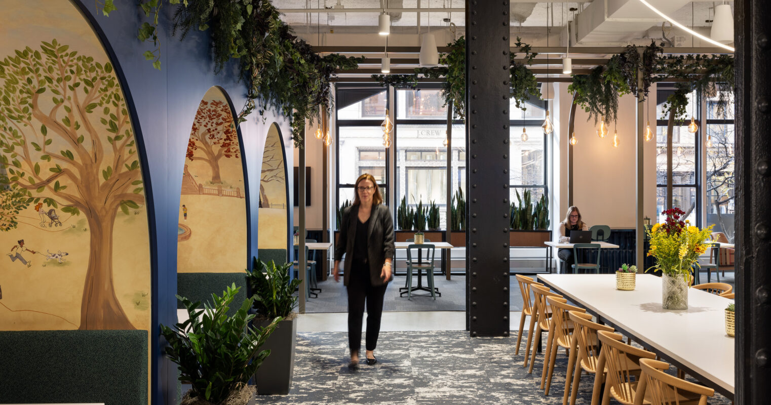 A spacious office interior featuring a blend of modern and natural elements. Arched murals of trees adorn the walls, complementing the vibrant greenery. A long communal table surrounded by wooden chairs fosters collaboration, while large windows provide abundant natural light and an inviting atmosphere for productivity.