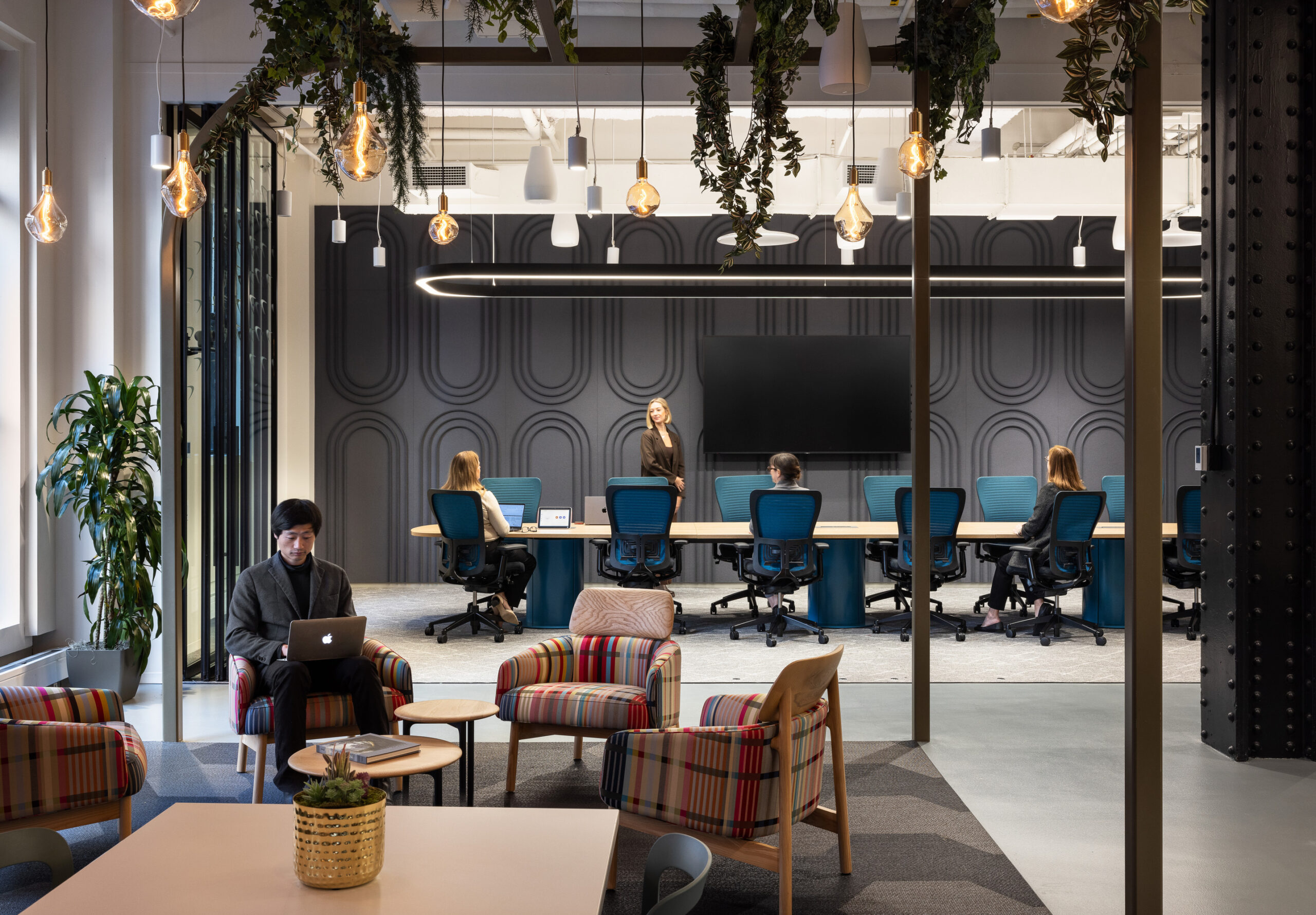 A modern office space featuring a collaborative environment with comfortable seating arrangements. The design includes a sleek conference room with a dark textured wall, equipped with a central screen. Natural light flows in, highlighting the greenery and stylish furniture, promoting a productive and inviting atmosphere for teamwork.