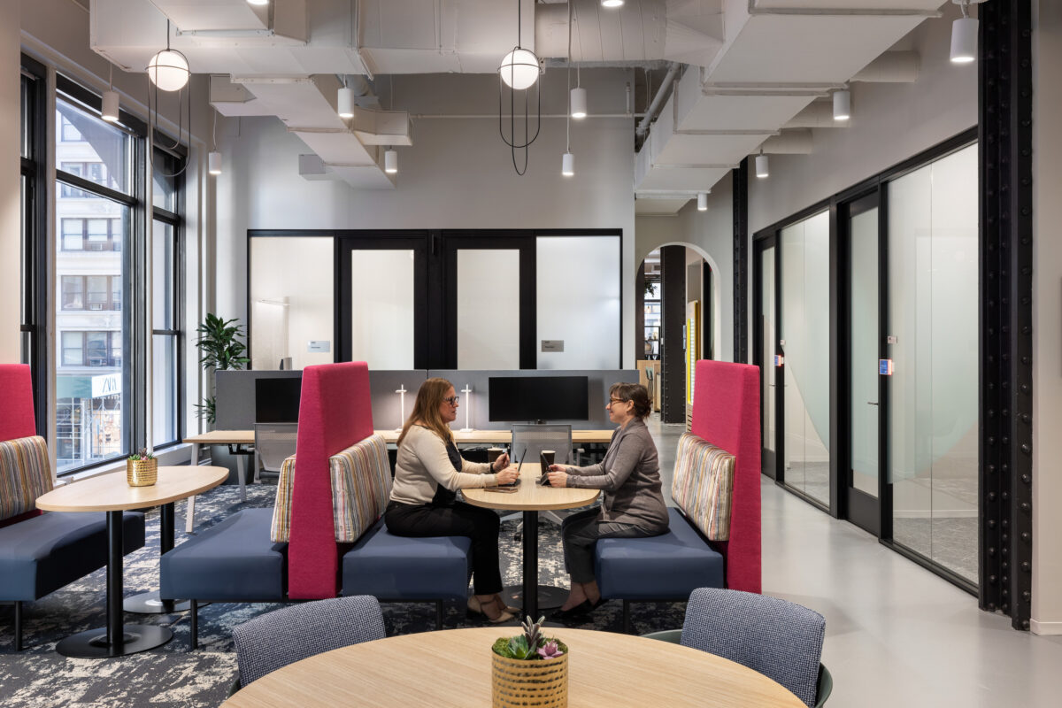 A modern office space featuring two individuals engaged in conversation at a rounded wooden table. The interior showcases subtle gray walls, accentuated by vibrant pink seating. Large windows provide natural light, enhancing the open layout. Stylish partitions and contemporary furnishings create a collaborative and inviting environment.