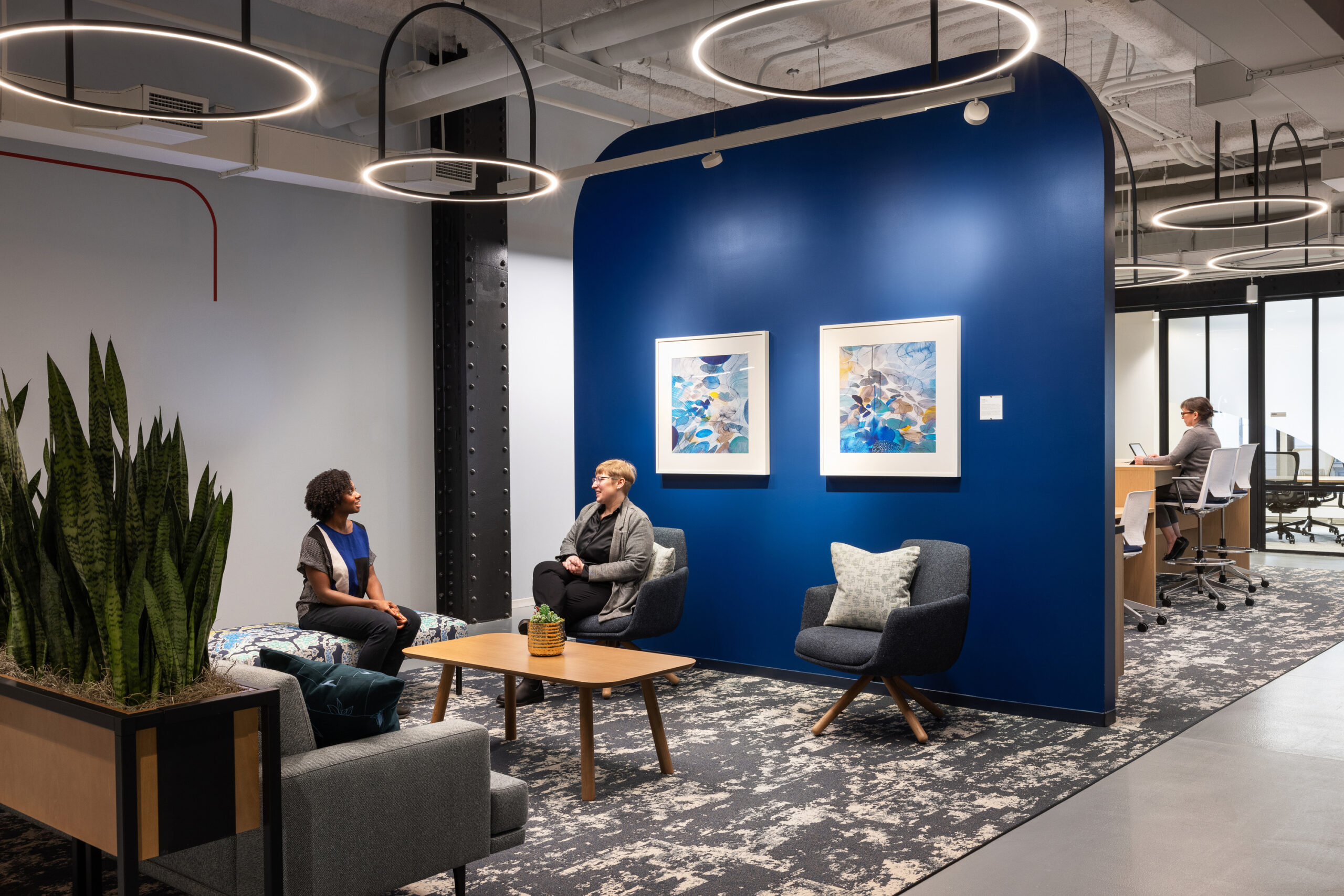 A modern office space featuring a bold blue accent wall, framed abstract artworks, and contemporary seating arrangements. Soft, sustainable textiles and greenery enhance the inviting atmosphere. Sleek pendant lighting adds an elegant touch, while a structured layout encourages collaboration and productivity among team members.