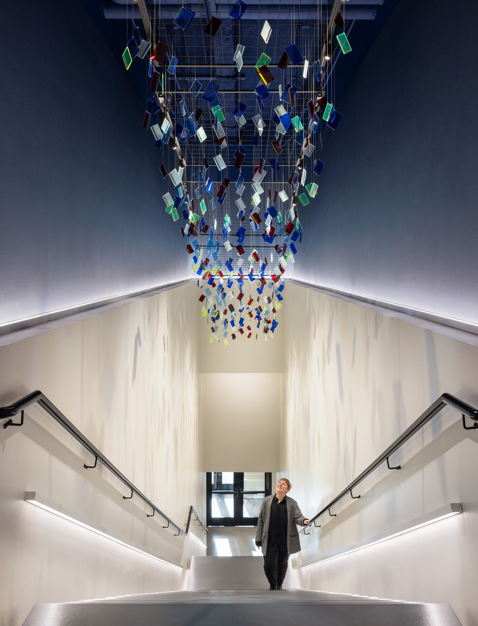 A modern staircase featuring sleek, minimalistic handrails and soft, ambient lighting. Above, a striking installation of colorful glass polygons suspended from the ceiling adds artistic flair and visual interest, creating a dynamic focal point that enhances the contemporary architectural space. The overall design emphasizes clean lines and vibrant color.