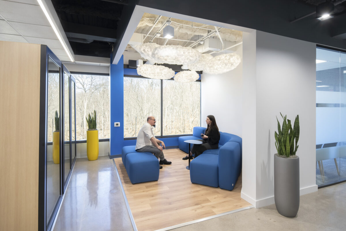 Contemporary office space featuring a cozy seating area with blue modular couches and a natural wooden floor. Large windows offer a scenic outdoor view, while suspended cloud-like light fixtures add an artistic touch. Green plants in sleek containers enhance the inviting atmosphere, encouraging collaboration and relaxation.