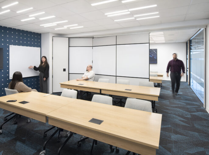 A modern conference room showcasing a collaborative workspace. Features include a series of light wooden tables arranged for group discussions, a dark blue accent wall with a geometric pattern, and large glass windows allowing natural light. Minimalistic design elements and technology-integrated tables enhance functionality and engagement.