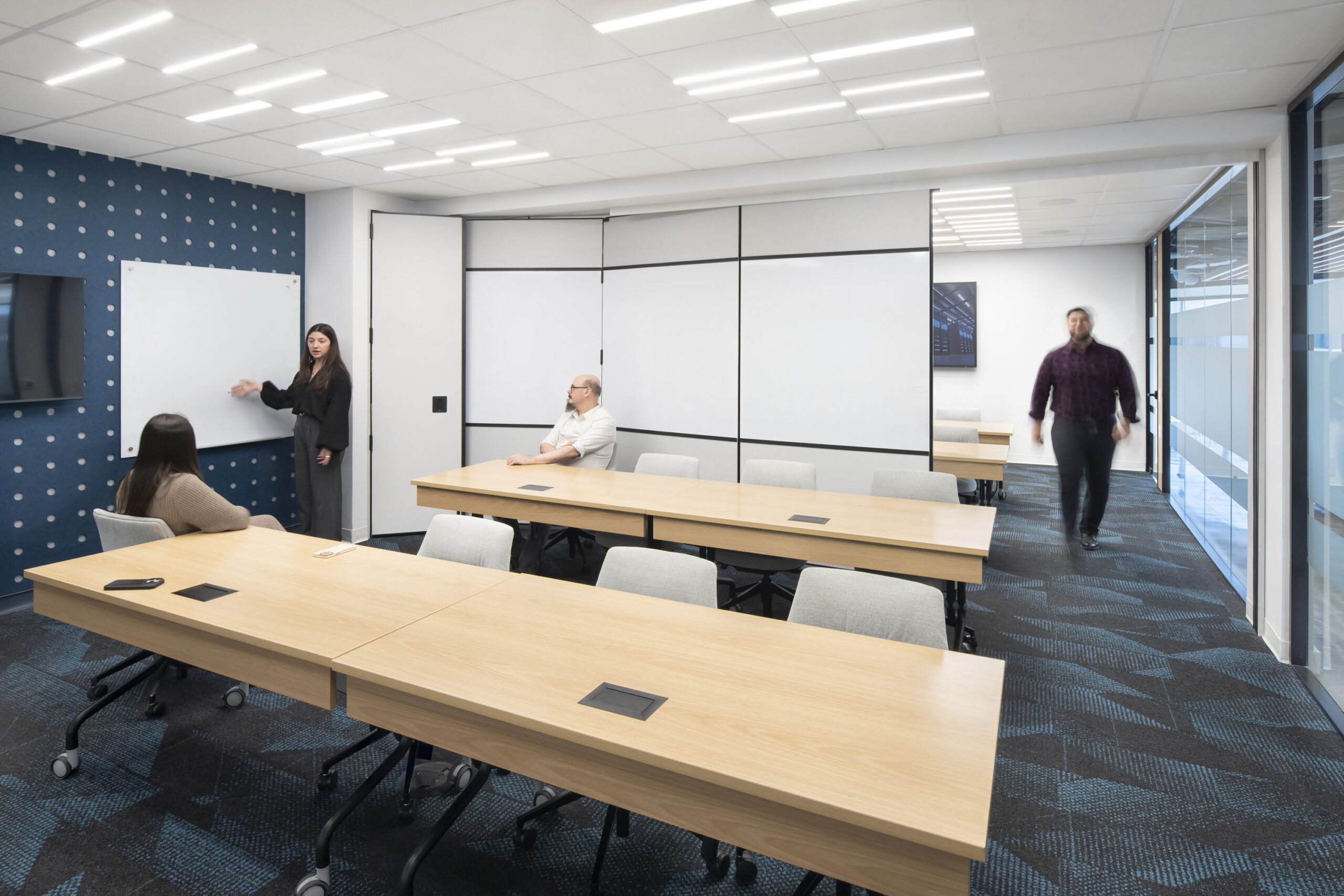 A modern conference room showcasing a collaborative workspace. Features include a series of light wooden tables arranged for group discussions, a dark blue accent wall with a geometric pattern, and large glass windows allowing natural light. Minimalistic design elements and technology-integrated tables enhance functionality and engagement.