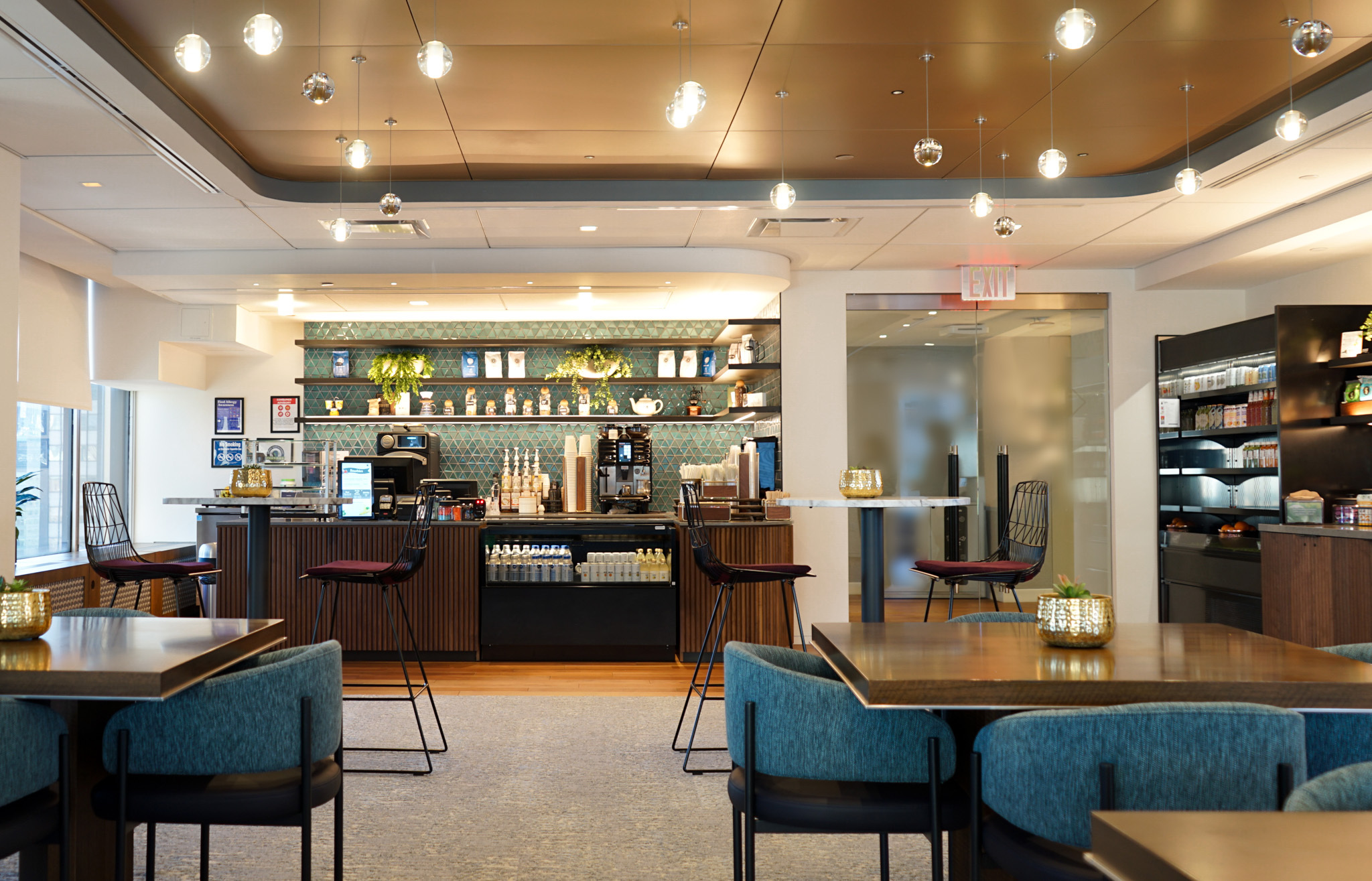A modern café interior features elegant turquoise seating and sleek wooden tables. The barista counter, adorned with geometric tile, offers a variety of beverages, enhanced by stylish light fixtures suspended from a gold-accented ceiling. Lush greenery accents complement the vibrant color palette, creating a welcoming atmosphere.