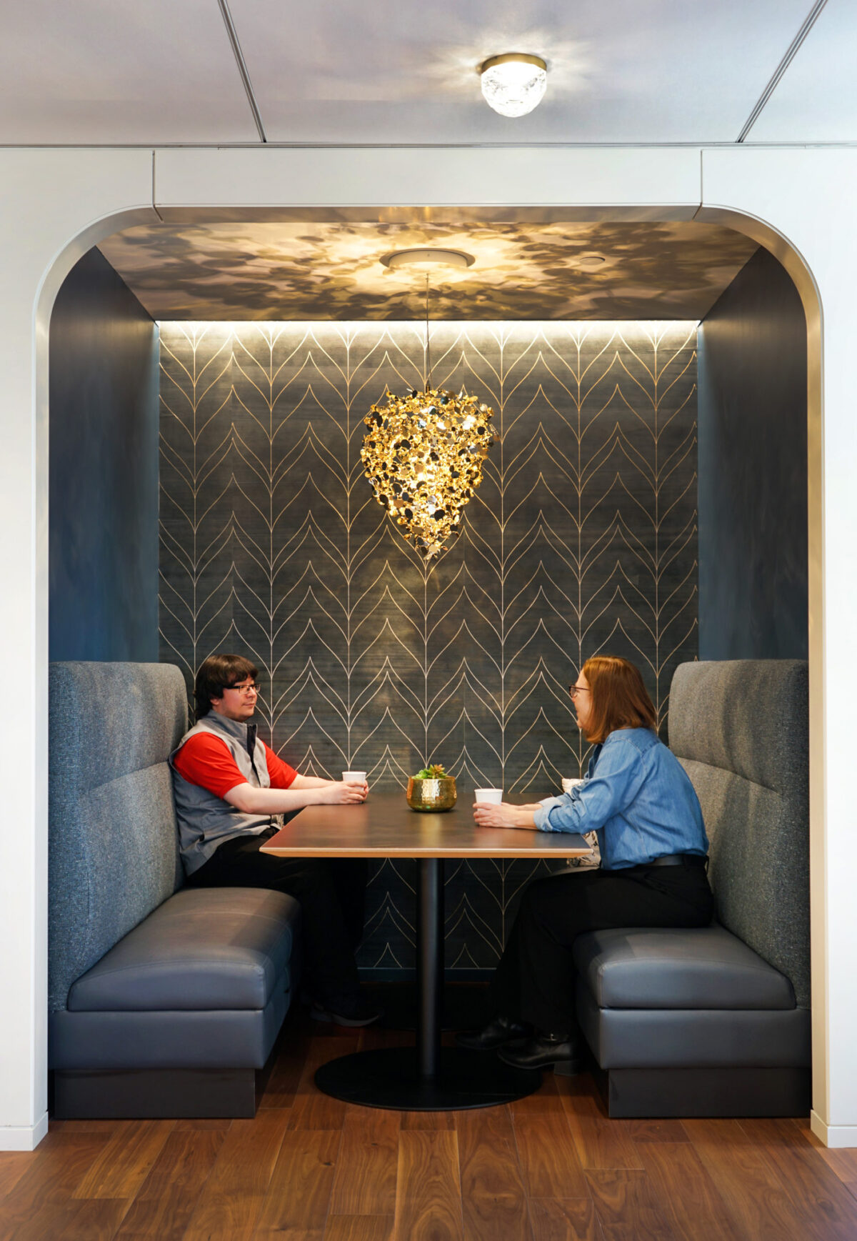 A modern meeting nook features sleek, upholstered booths with high backs, providing an intimate setting for conversation. Soft lighting emanates from a decorative pendant light designed to resemble foliage, while a patterned accent wall enhances the atmosphere with texture and sophistication, promoting creativity and collaboration.