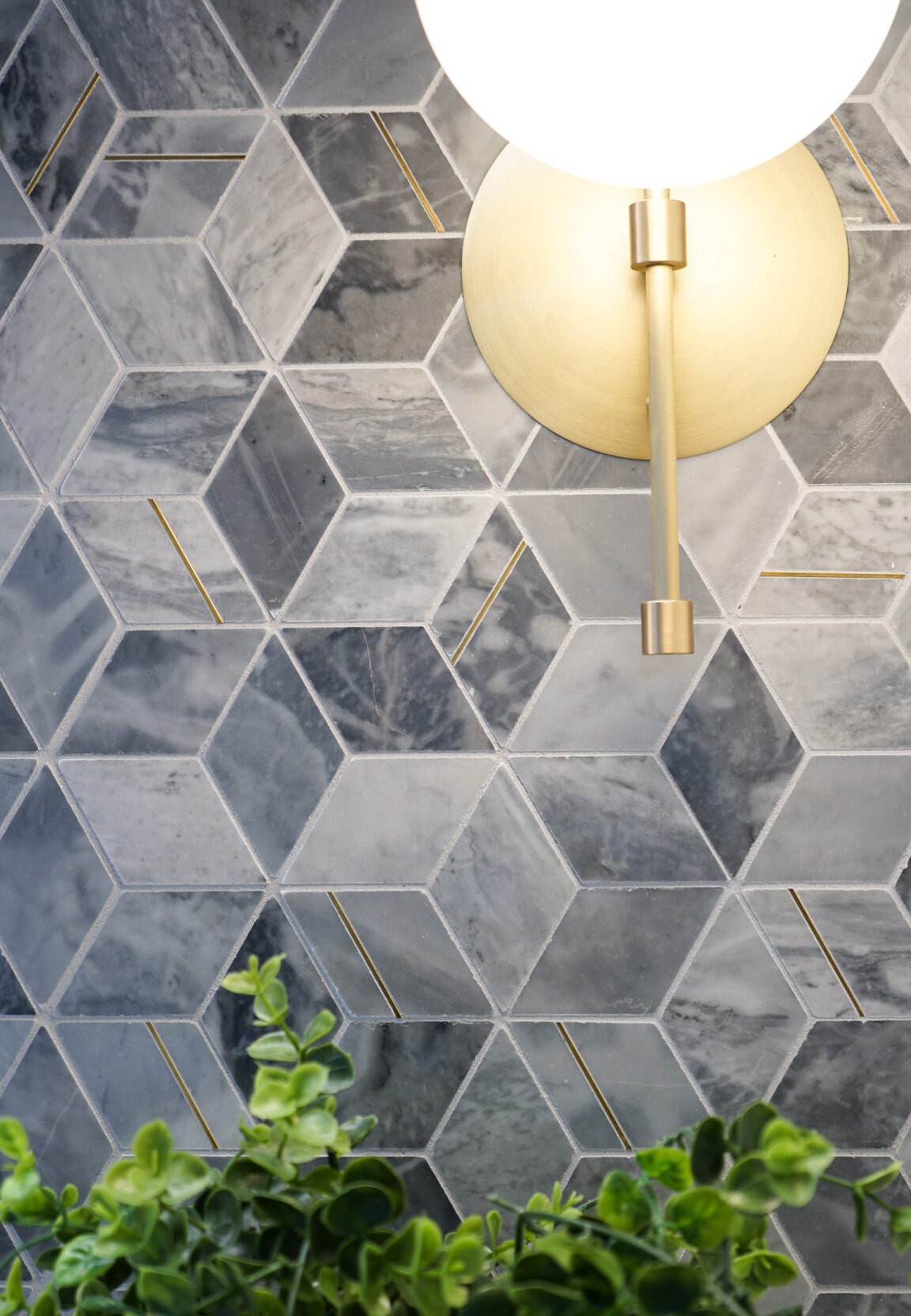 A modern hexagonal tile backsplash features a blend of cool gray tones, accented with thin gold strips. A sleek, gold wall sconce casts warm light, creating an inviting atmosphere. Beneath, lush greenery adds a touch of nature, enhancing the sophisticated design with organic elements.