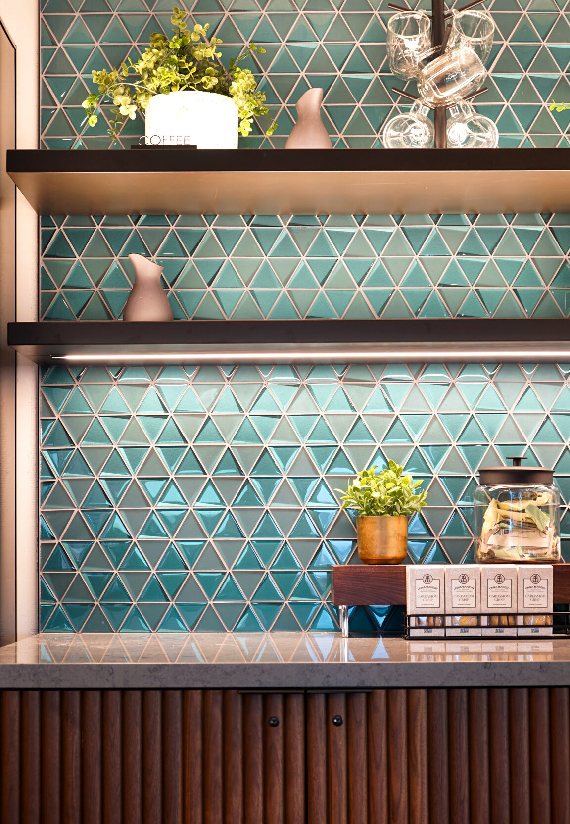 Textured teal tiles create a striking geometric pattern on the backsplash, contrasted by minimalist wooden shelves displaying ceramic vases and greenery. Below, a sleek wooden cabinet with vertical slats houses neatly organized coffee-related items, integrating functionality with contemporary design aesthetics in a chic interior space.