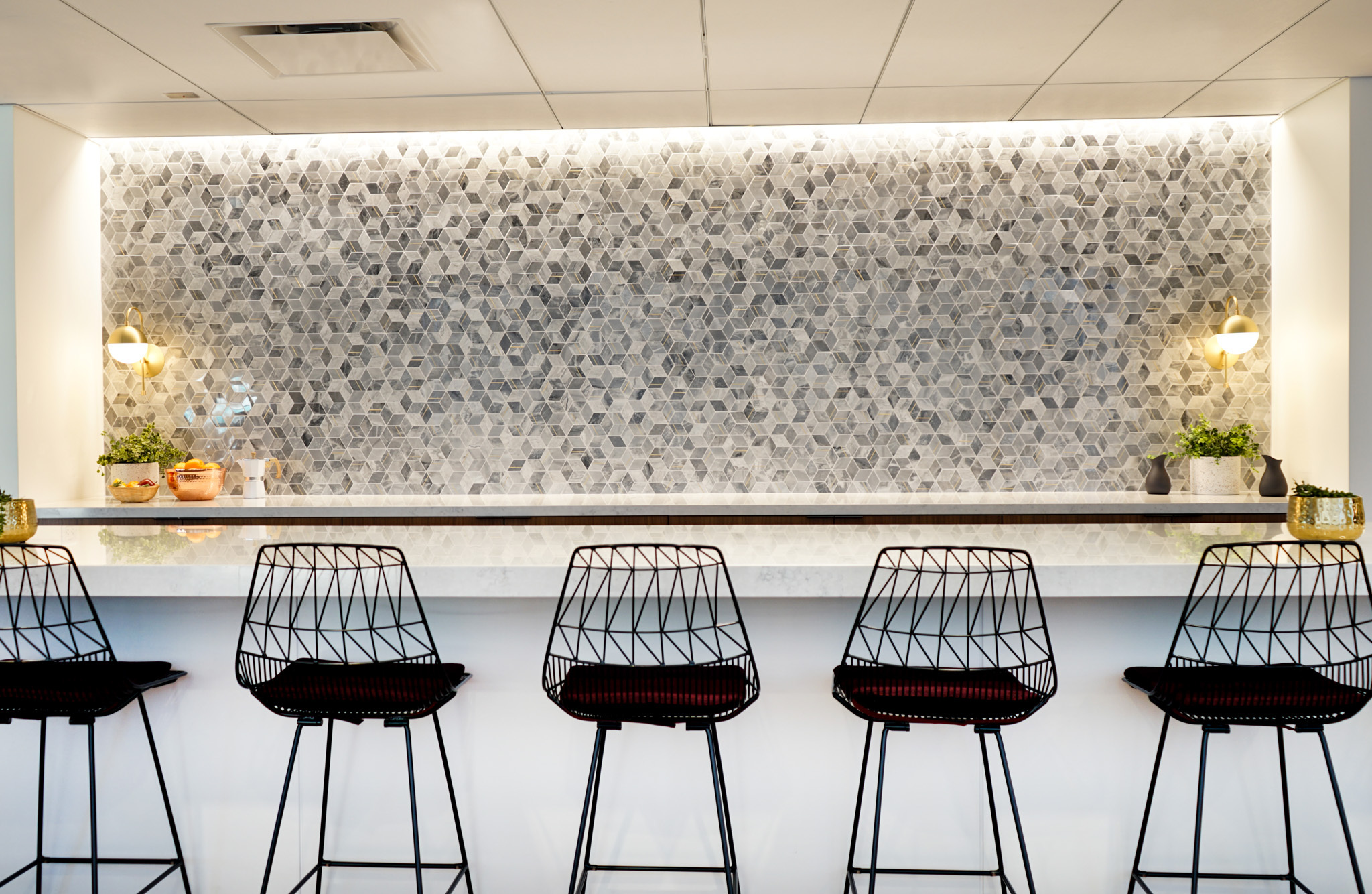 A sleek modern bar area features a white marble countertop paired with black wire-frame stools. The backdrop showcases a striking mosaic wall made of varying shades of gray stone, accentuated by warm, ambient lighting from wall-mounted fixtures. Small potted plants add a touch of greenery.