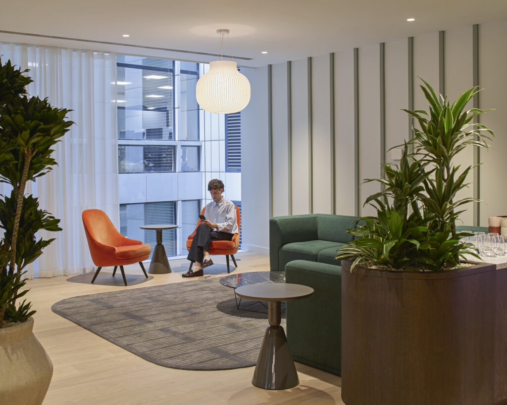 A modern lounge featuring a harmonious blend of colors and textures. Cozy orange chairs complement a green sectional sofa, while a geometric rug anchors the space. Lush indoor plants enhance the inviting atmosphere, illuminated by a distinctive pendant light, creating an aesthetically pleasing and functional environment for relaxation and conversation.