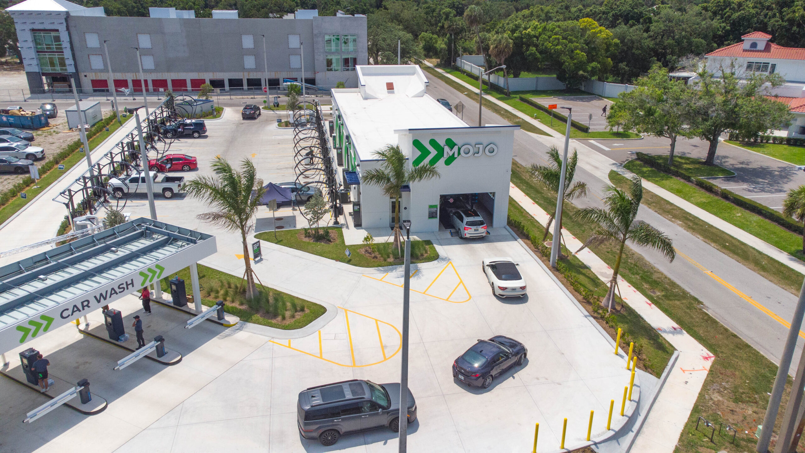 A modern car wash facility features a minimalist architectural design with a bright white exterior and bold green branding. The layout includes several car washing stations, palm trees for landscaping, and designated parking areas. Enhanced with efficient elements, the design prioritizes both functionality and aesthetic appeal.