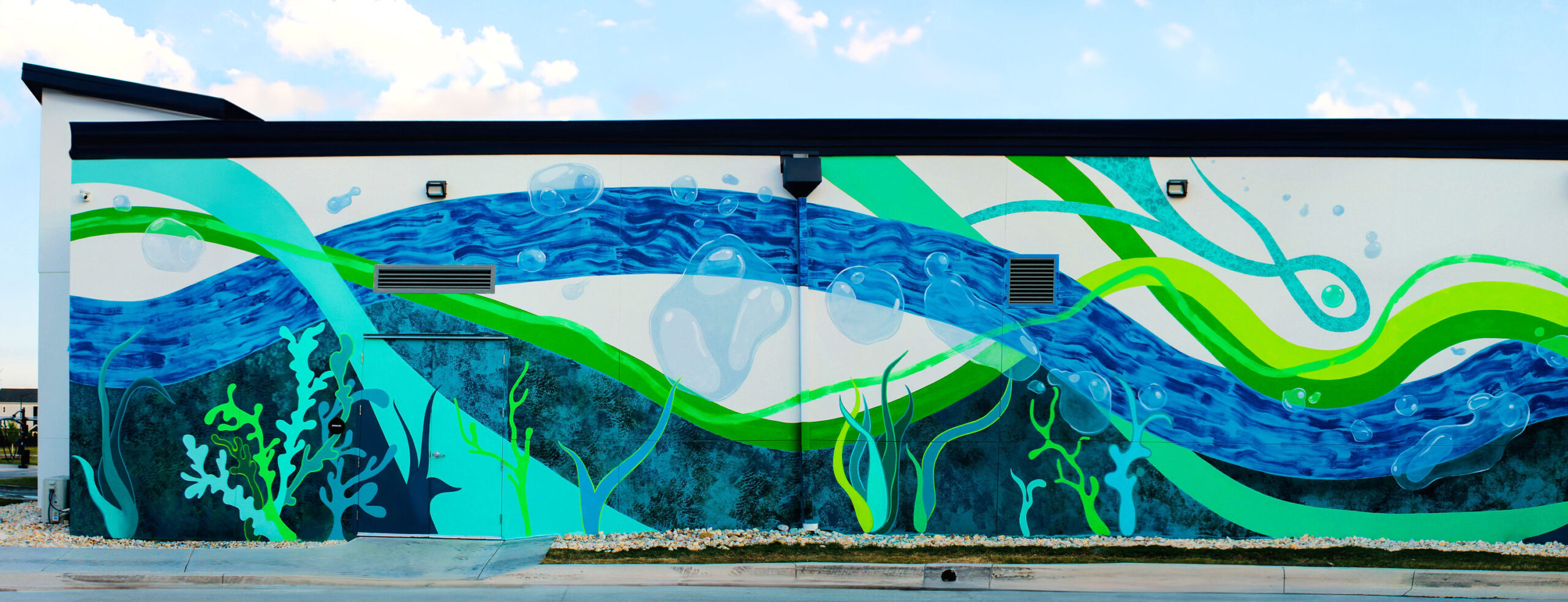 Vibrant mural showcasing fluid, wavy patterns in shades of blue and green, mimicking underwater currents. The artwork features stylized bubbles and coral-like structures, creating a lively aquatic theme. This striking design enhances the exterior of the building, fostering a sense of creativity and connection to nature in the urban landscape.