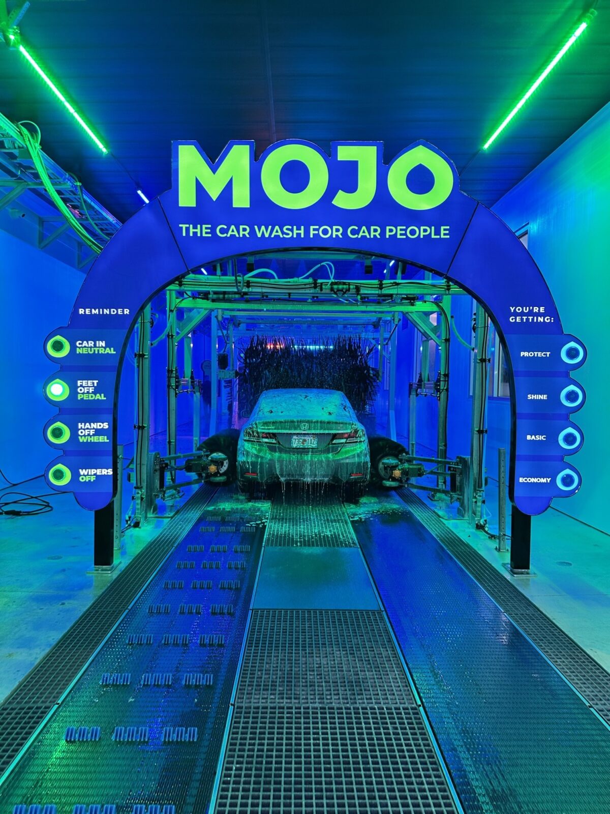 A vibrant car wash facility featuring a bold blue and green color scheme, highlighting the "MOJO" sign. The illuminated archway leads to a modern automated washing bay, where a vehicle is being cleaned. Informative signage outlines operational guidelines and service features, enhancing user experience and engagement.