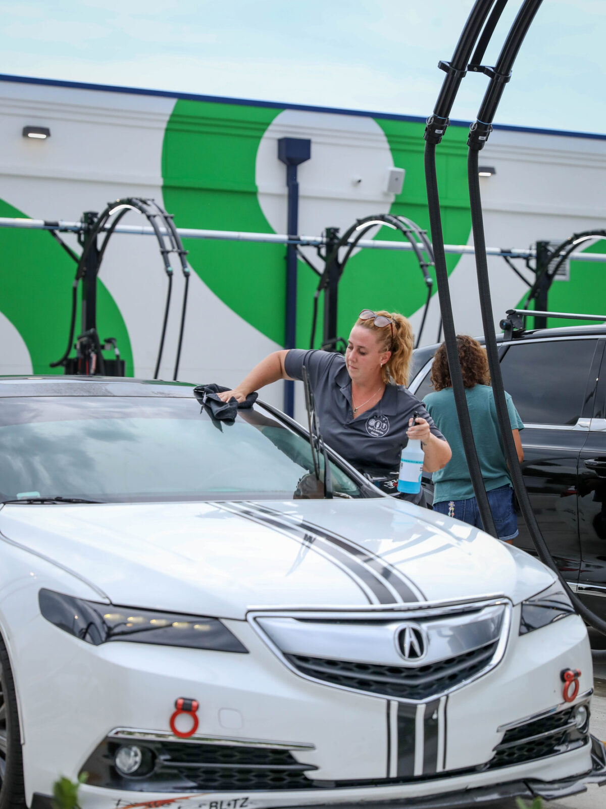 A worker meticulously cleans the windshield of a modern white sedan, featuring sleek black racing stripes. Surrounding vehicles and a vibrant green and white mural create an energetic atmosphere at the car wash. Attention to detail highlights the professionalism in vehicle maintenance and care within this lively environment.