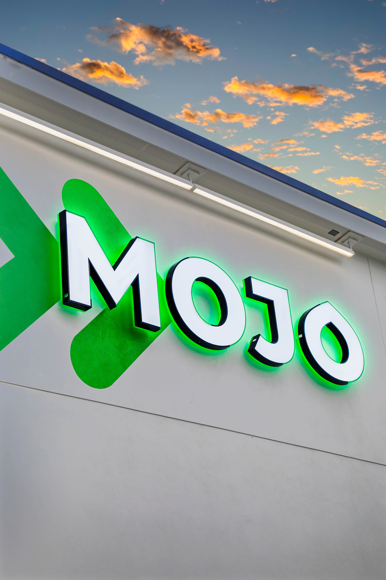 The façade features the bold "MOJO" signage, illuminated with a vibrant green glow that contrasts against the clean, white background. Geometric shapes accompany the text, enhancing the modern aesthetic. Natural lighting showcases the subtle texture of the wall, creating an inviting and contemporary appearance.
