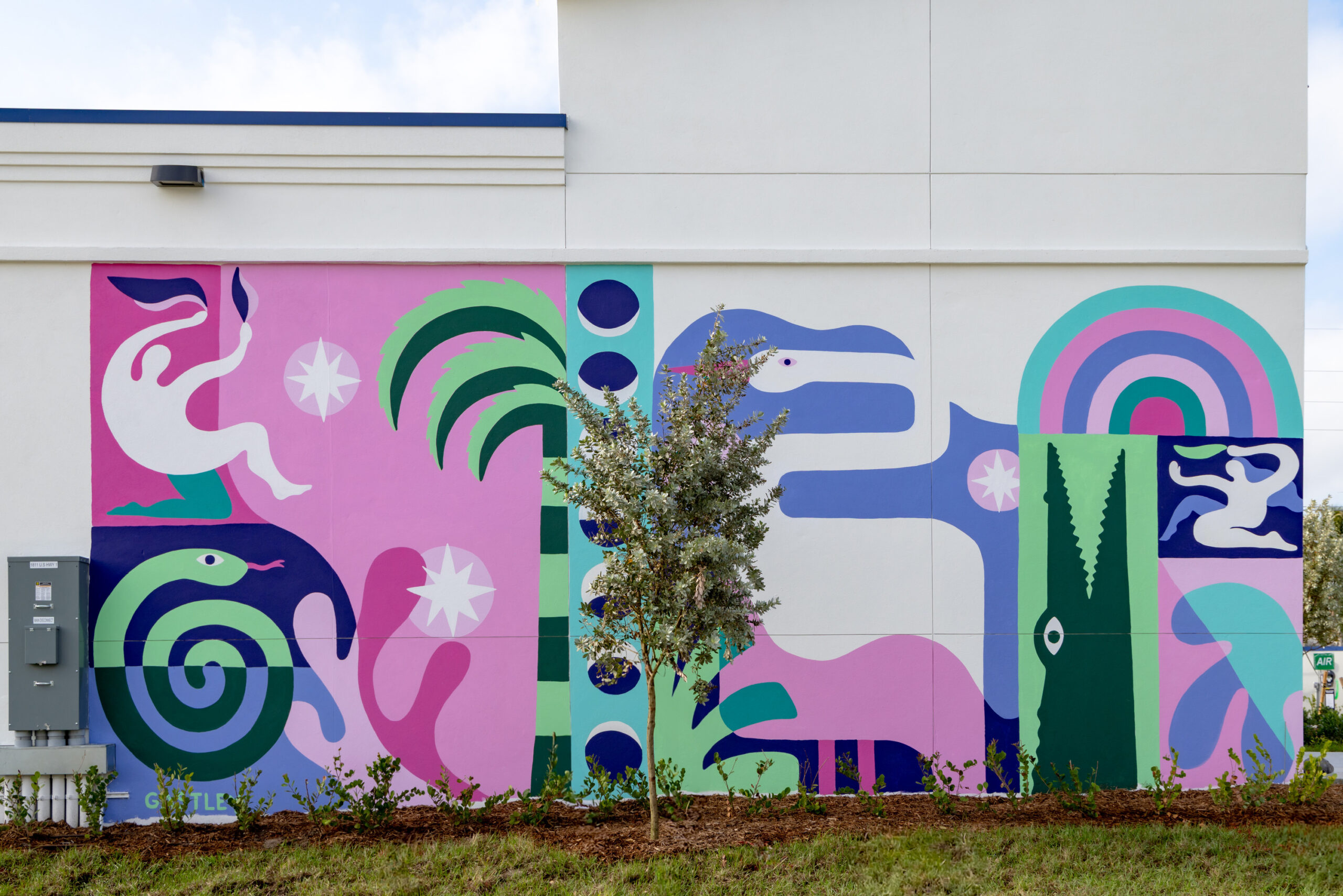 A vibrant mural adorns the exterior wall, featuring playful shapes and bold colors, including greens, pinks, and blues. Stylized figures, abstract patterns, and whimsical elements like palm trees and serpents create a lively atmosphere, enhancing the architectural context while promoting community engagement through artful expression.